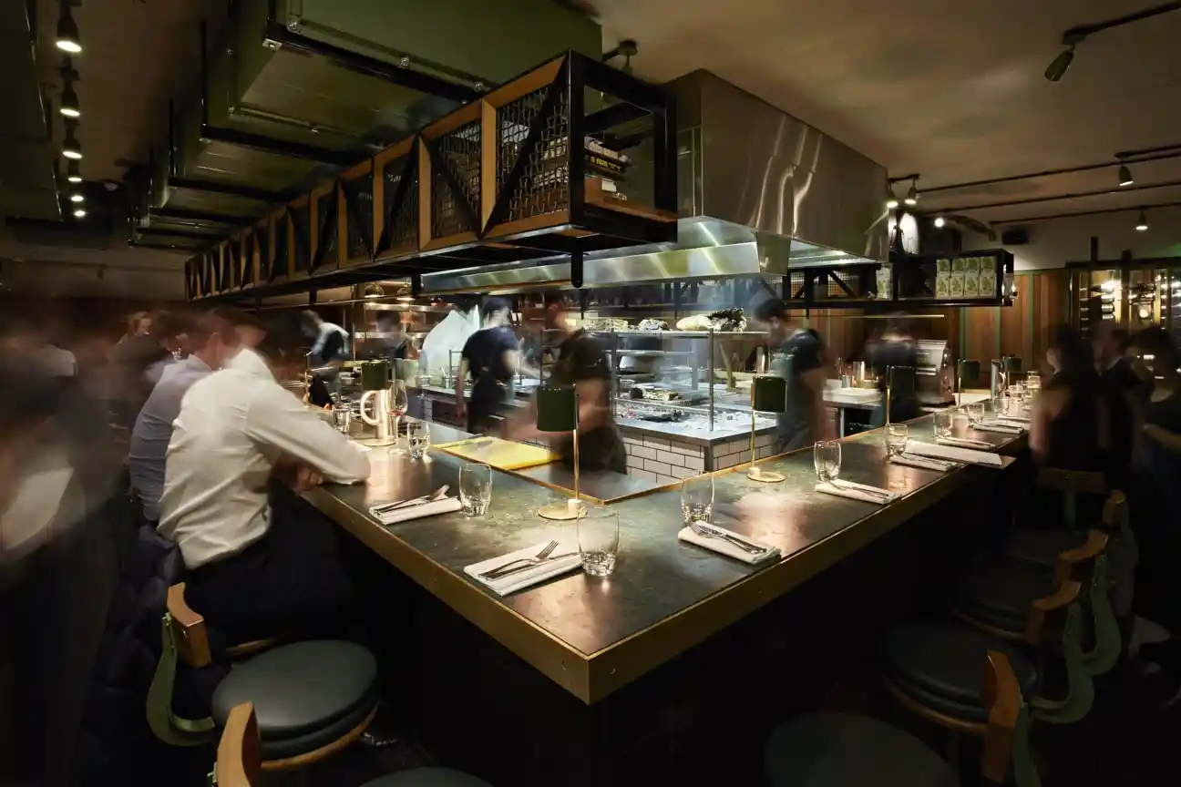 Diners seated at the counter of a London steakhouse, enjoying fire-cooked steaks and cocktails in a lively setting.